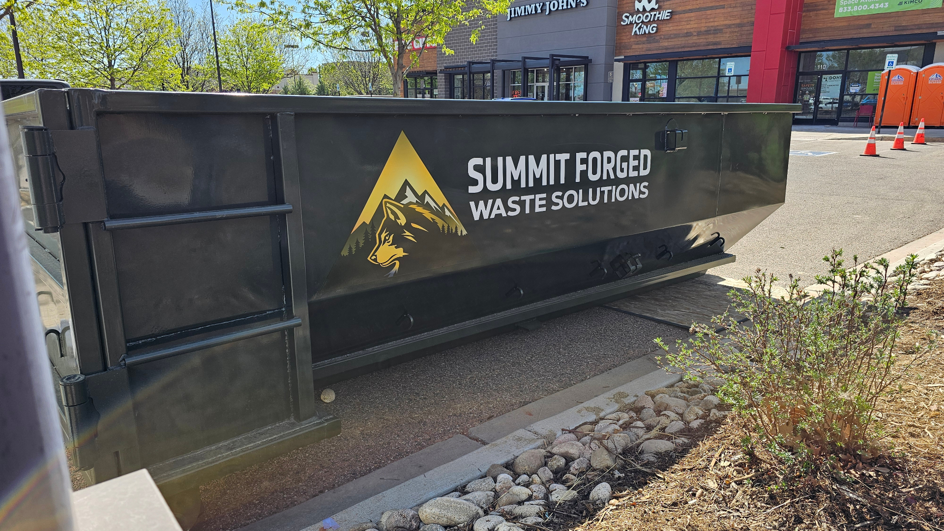 Summit Forged branded roll-off dumpster on the job at a Northern Colorado shopping center, showing the deep olive container and gold wolf logo